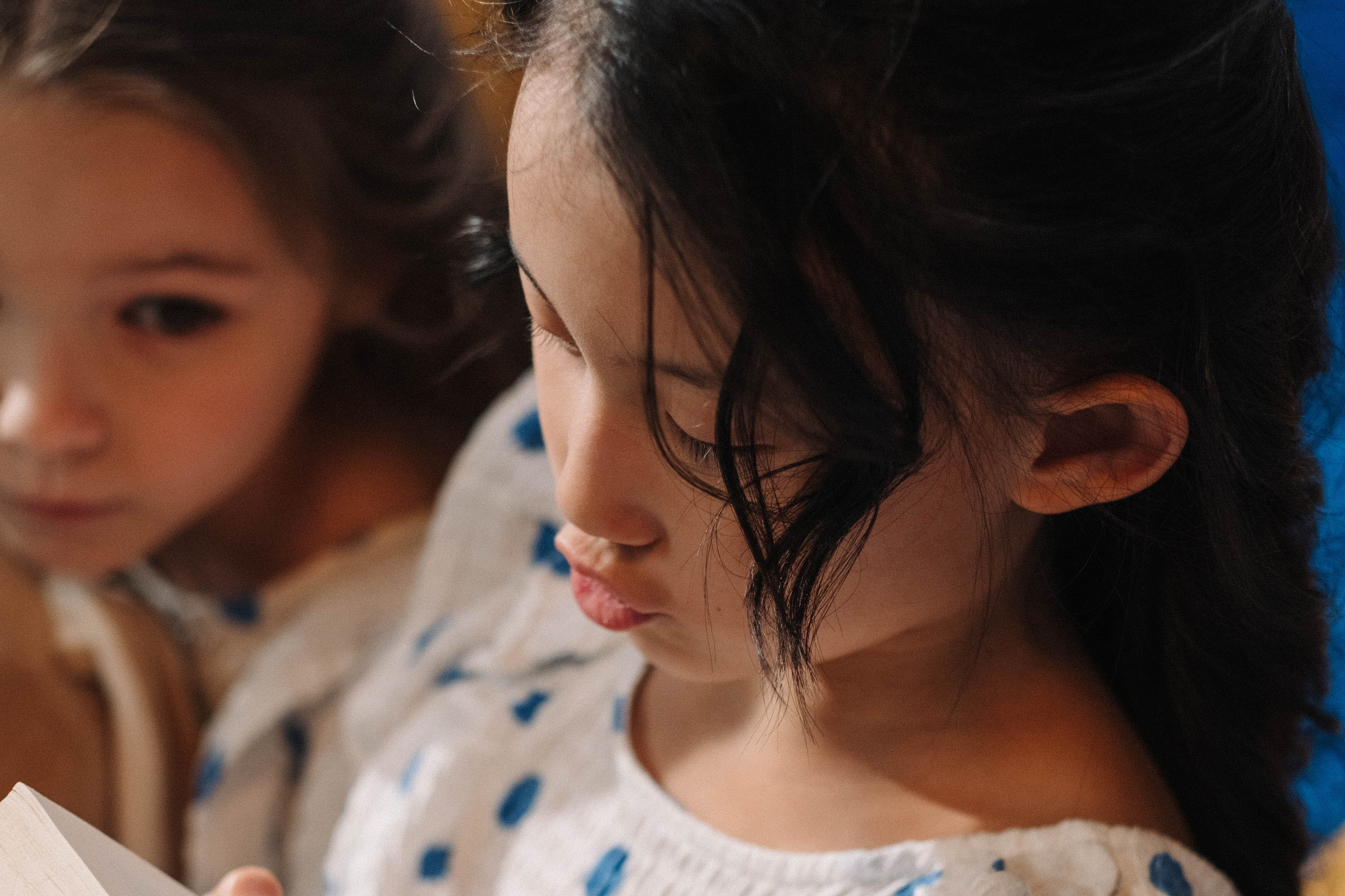 Image: two girls in polka dot dresses sit closely, with one girl intently reading a book.